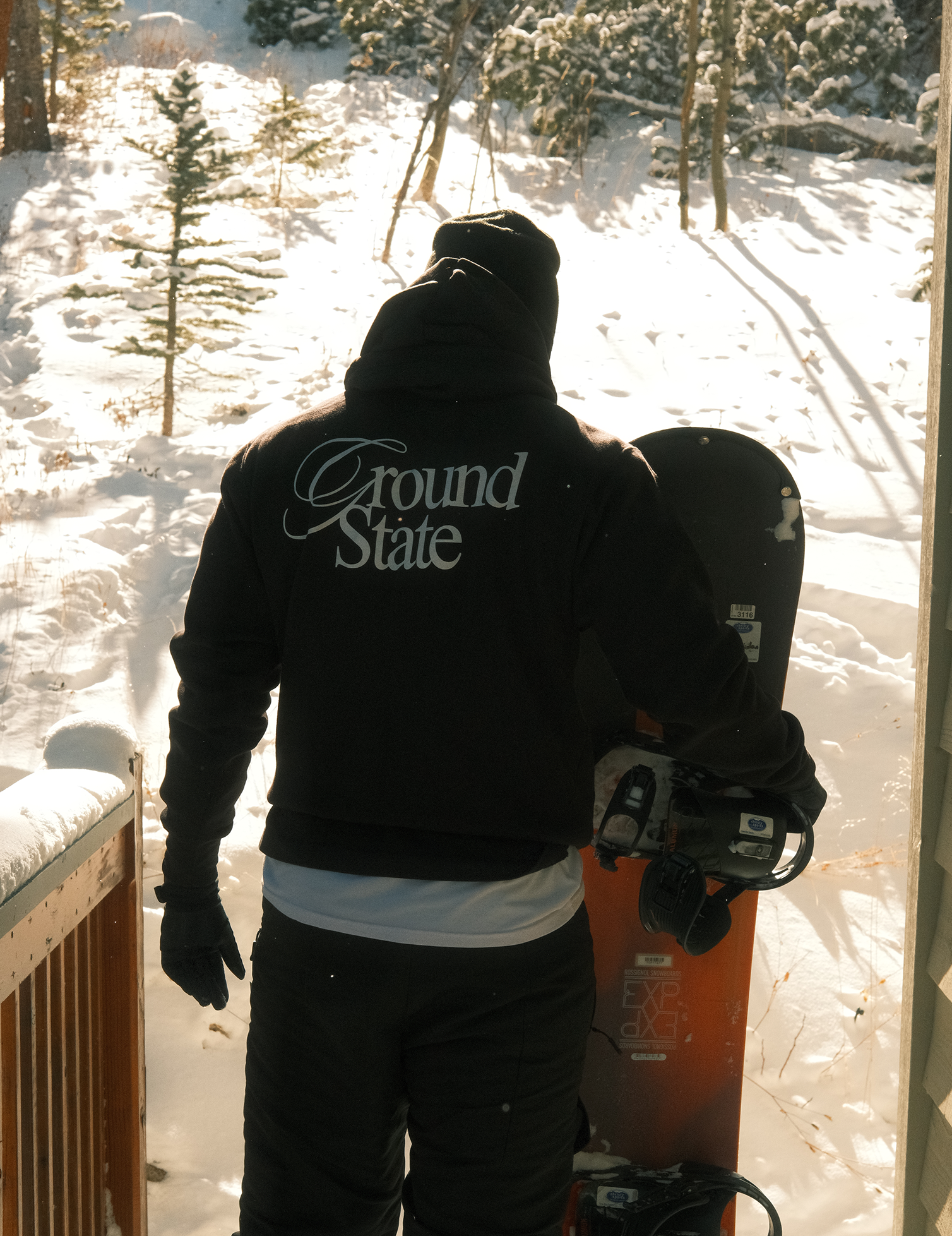 Person holding a snowboard with 'Ground State' branding, standing on a wooden deck with snow-covered ground and trees in the background.